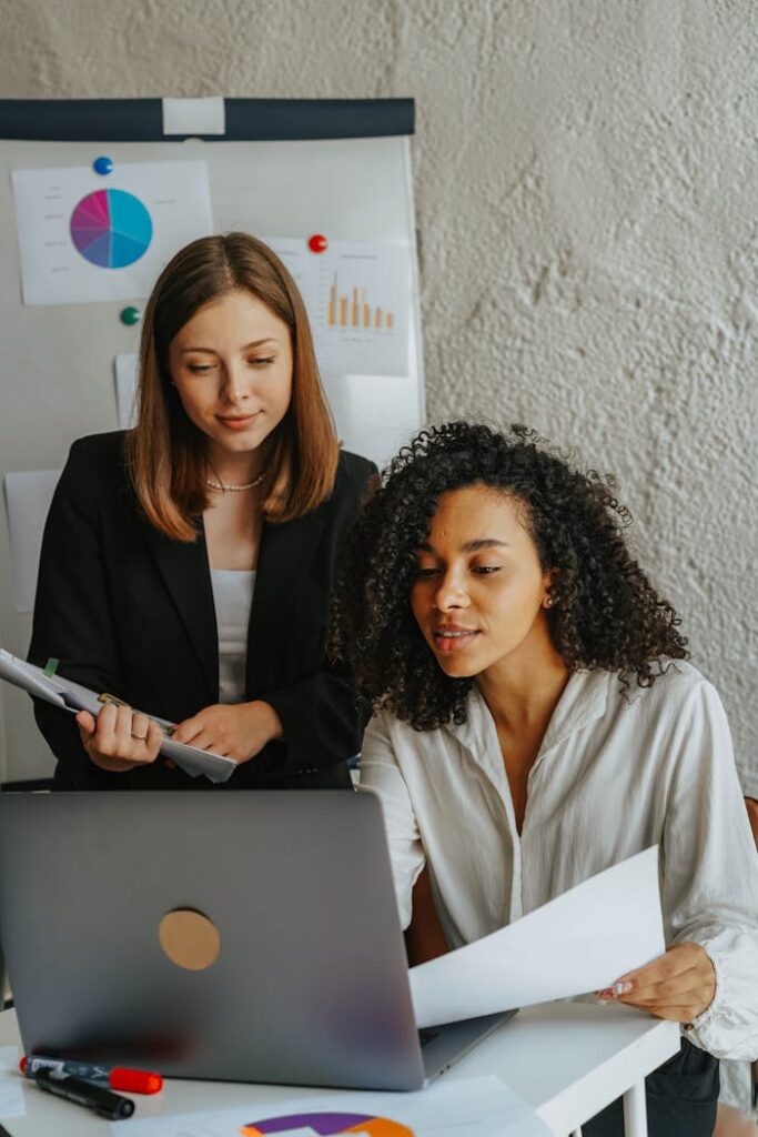 Two Women Holding Papers while Looking at Laptop Screen